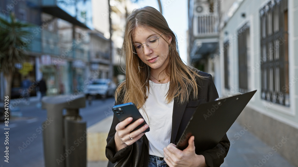 Fototapeta premium A young blonde woman in business attire checks her phone while holding a clipboard on a busy city street.
