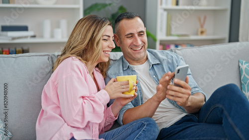 Man and woman couple drinking coffee using smartphone sitting on sofa at home