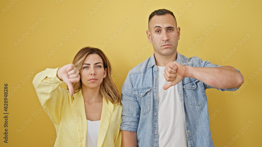 Man and woman couple standing with serious face doing thumb down gesture over isolated yellow background Man and woman couple standing with serious face doing thumb down gesture over isolated yellow background
