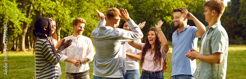 Group of a happy young people having fun giving each other high five while walking in the summer park. Cheerful excited multiethnic friends laughing outdoors. Teamwork and friendship concept.