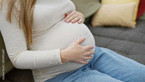 Pregnant hispanic woman caressing her belly, seated on a couch indoors with cushions in the background.
