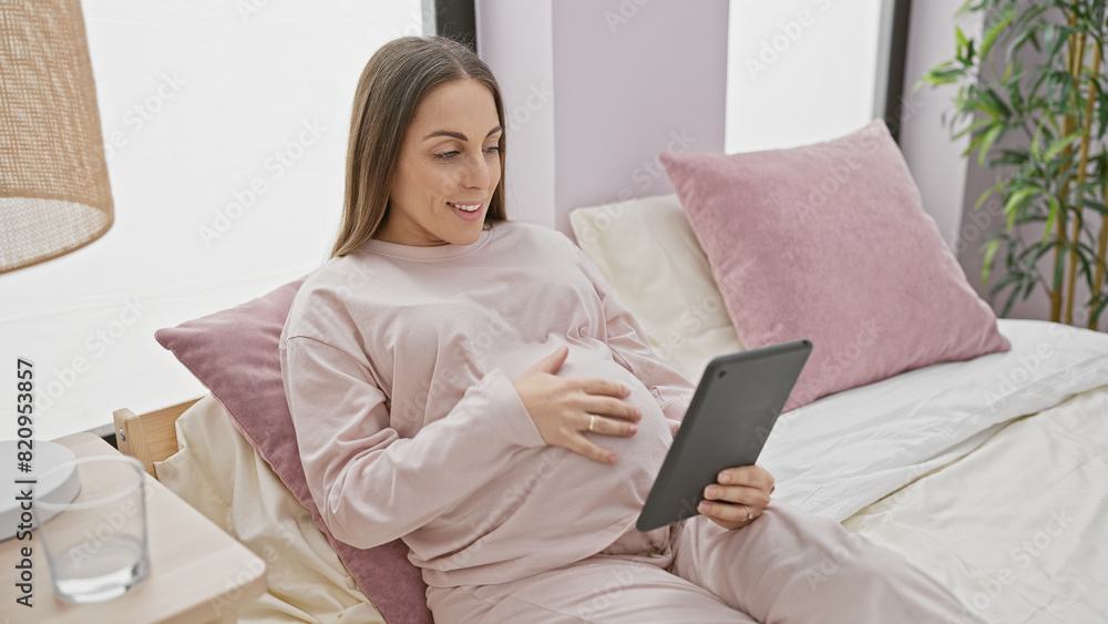 Hispanic pregnant woman smiles while holding a tablet, comfortably resting in a home bedroom setting. Hispanic pregnant woman smiles while holding a tablet, comfortably resting in a home bedroom setting.