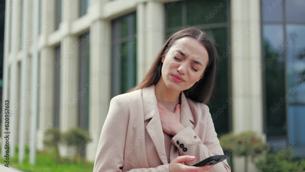 Mournful caucasian female feeling sharp pain in chest while scrolling websites on wireless smartphone outdoors. Young woman looking around and searching help on background of office corporate.