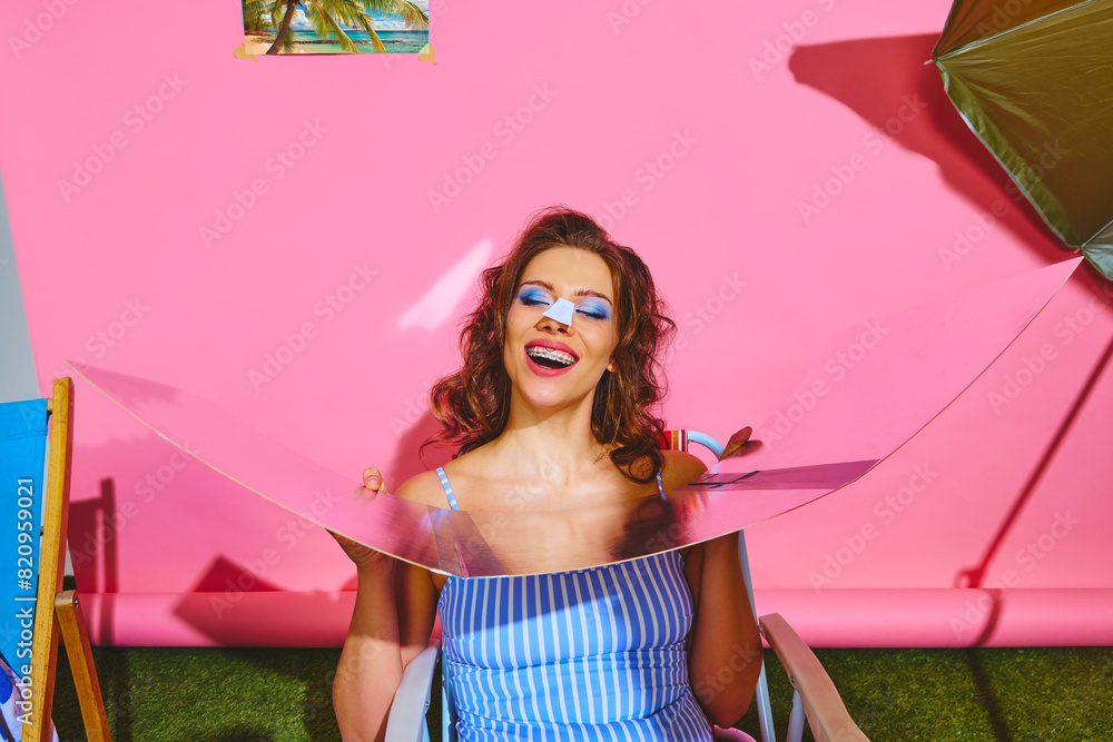 Fototapeta premium Portrait of overjoyed, smiling woman lounges sitting against pink background with picture of beach. Summery atmosphere. Concept of pop art, remote work, travelling, party, recreation, lifestyle, style