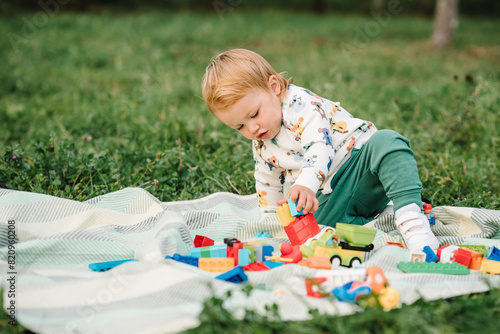 Little cute toddler boy walking and playing small car toys in garden. Child 2 year old plays constructor game sitting in green grass on blanket in park sunny summer day. Outdoors creative activities.