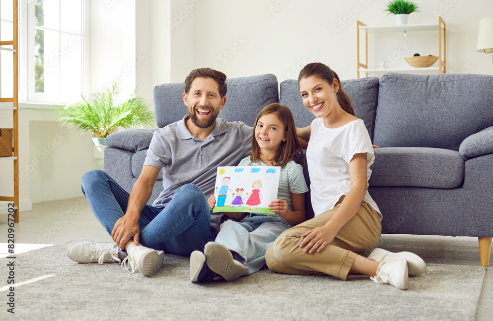Smiling child girl sitting on the floor at home showing drawing of a happy family. Portrait of joyful parents with their cute daughter looking cheerful at camera. Family leisure concept. Smiling child girl sitting on the floor at home showing drawing of a happy family. Portrait of joyful parents with their cute daughter looking cheerful at camera. Family leisure concept.