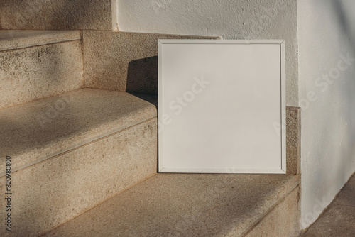Closeup of empty square white thin picture frame leaning against wall in sunlight. Outdoor sandstone stairs background, shadows overlay. Poster mockup for art display. Minimal summer design. No people