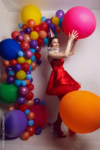 Funny clown woman in a red dress and a hat stands against a background of colored balloons