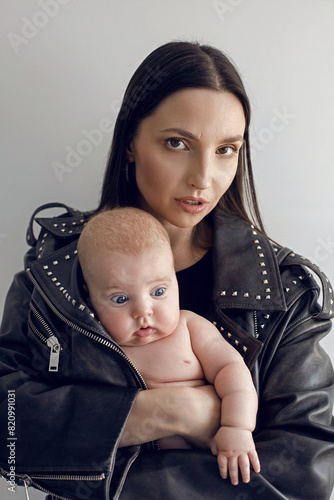 Happy mother rocker with a baby daughter stands in a leather jacket with rivets against a white wall in the studio