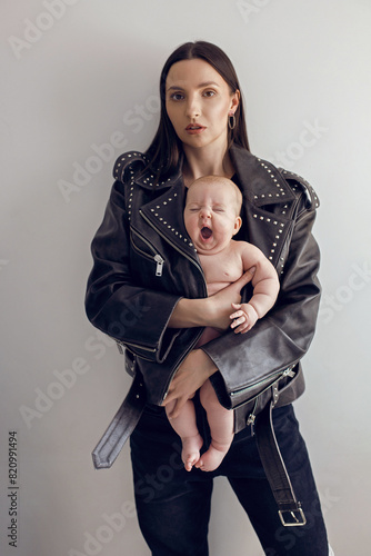 Happy mother rocker with a baby daughter stands in a leather jacket with rivets against a white wall in the studio. yawn.