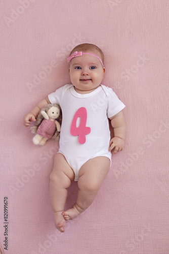girl baby infant with a bow lies on the bed in a white bodysuit with a doll toy next to it, the child has a pink number four months on a pink background