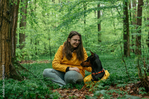 young woman walking with her doberman dog in the forest. Wearing yellow rain jackets girl and dog playing outdoors. Human and dog friendship concept
