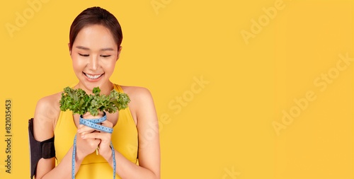 Smiling young woman in a yellow sportswear holding a bunch of fresh vegetable with a measuring tape, symbolizing healthy eating and fitness Studio shot and copy space Health care and wellness concept
