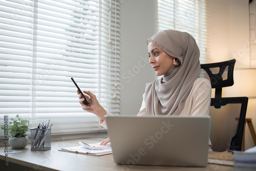 Muslim woman using smartphone at office desk. Professional workspace, casual attire