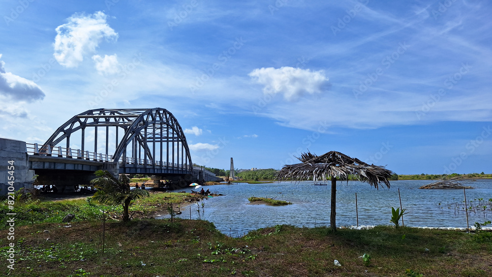 Obraz premium View of the estuary towards the sea with a bridge and a mangrove forest conservation area with a cloudy blue sky.