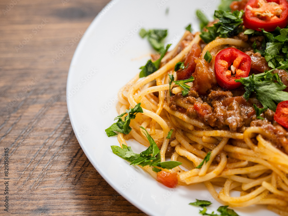 A plate of spaghetti with meat sauce and peppers. The dish is topped with a generous amount of red peppers and parsley. Italian style food on wooden table.