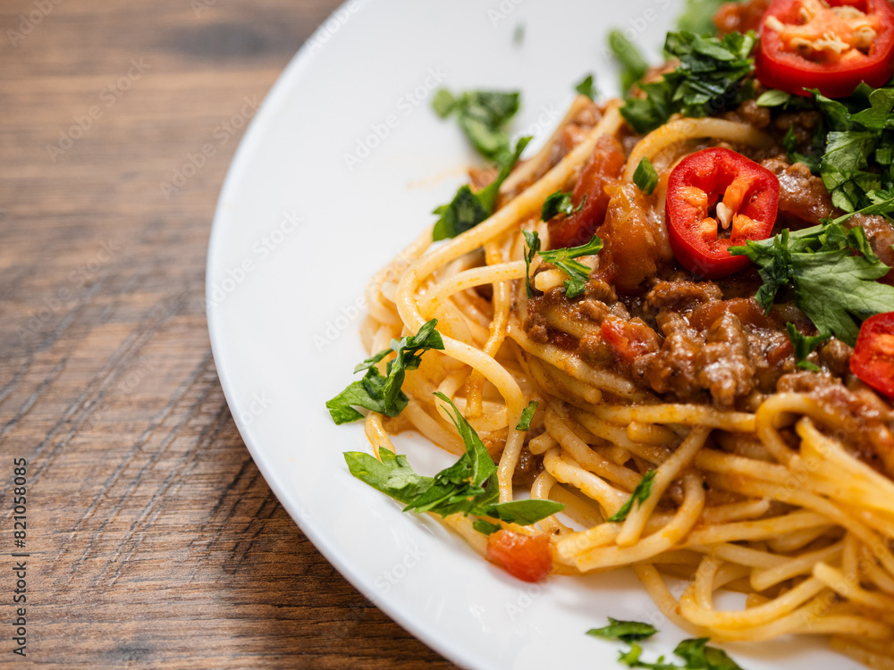 A plate of spaghetti with meat sauce and peppers. The dish is topped with a generous amount of red peppers and parsley. Italian style food on wooden table. A plate of spaghetti with meat sauce and peppers. The dish is topped with a generous amount of red peppers and parsley. Italian style food on wooden table.