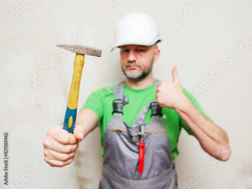 A man in a green shirt and gray overalls holding hummer in focus. Male construction worker with grey beard in white safety plastic helmet. Light color background.