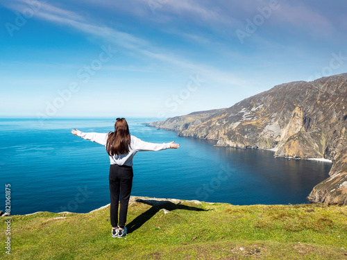 Teenager girl on a trip to Slieve League Cliff, Ireland. Stunning nature scenery with cliff, ocean and clean blue sky. Travel and tourism concept. Model posing in nature scene. Sunny day.