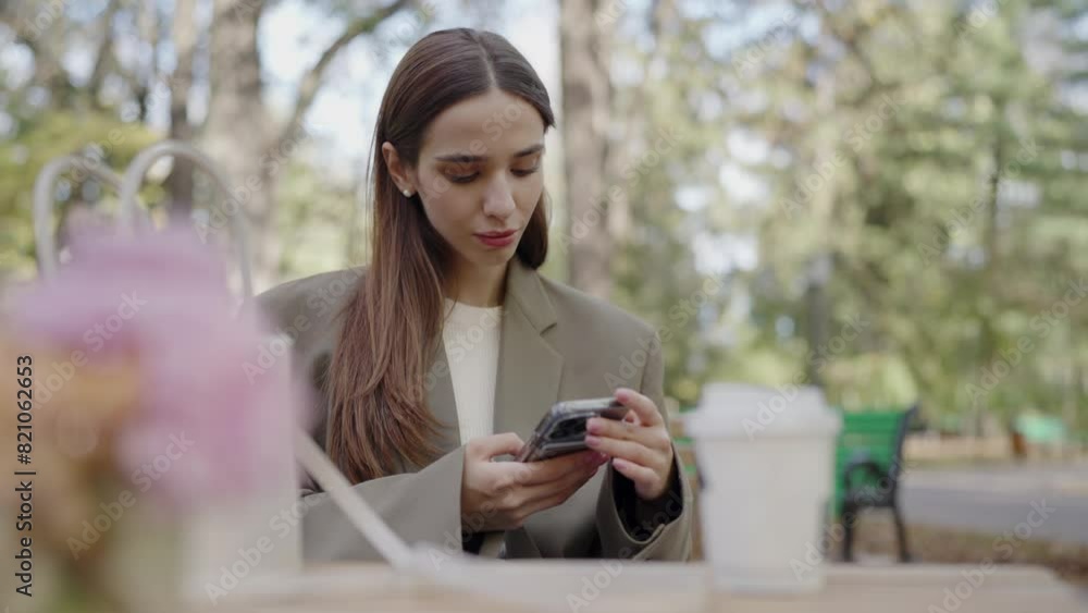 A young professional woman, dressed in a beige blazer, focusing intently on her smartphone while sitting at a park table. Young Professional Woman Texting on Smartphone in City Park A young professional woman, dressed in a beige blazer, focusing intently on her smartphone while sitting at a park table. Young Professional Woman Texting on Smartphone in City Park