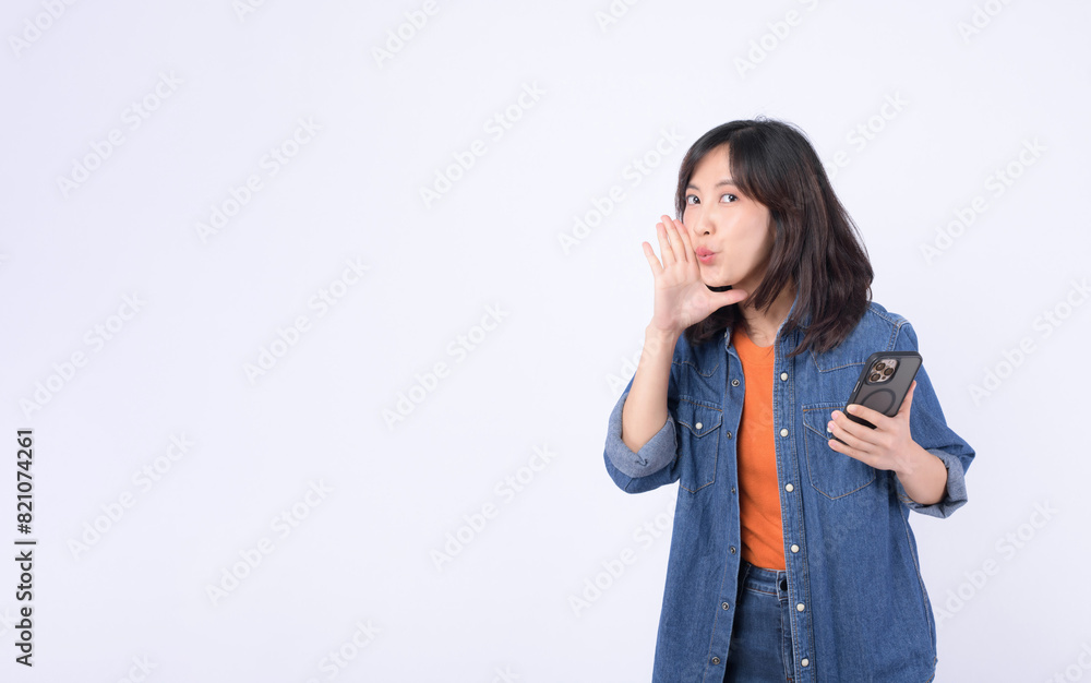 Asian woman wearing orange t shirt and denim jean is posing with a shouting gesture while holding smartphone against a white background. Asian woman wearing orange t shirt and denim jean is posing with a shouting gesture while holding smartphone against a white background.