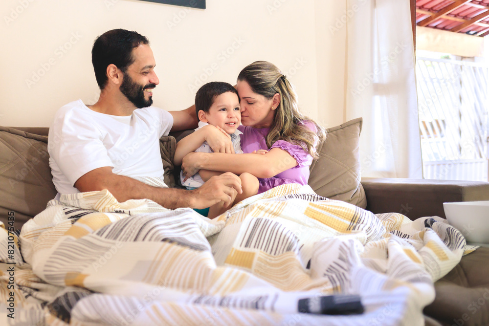 Father and mother hugging their young son on the couch at home in a gesture of love and affection. They are watching a movie through a streaming service and enjoying the moment together as a famy. Father and mother hugging their young son on the couch at home in a gesture of love and affection. They are watching a movie through a streaming service and enjoying the moment together as a famy.
