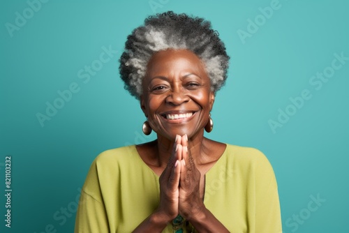 Portrait of a grinning afro-american elderly woman in her 90s joining palms in a gesture of gratitude isolated on solid color backdrop