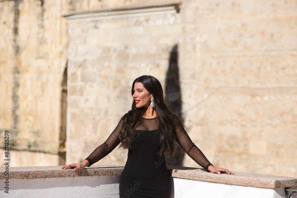 Beautiful young Spanish brunette woman with make-up and dressed in an elegant black dress. The woman is leaning on the railing. Beauty and fashion concept.