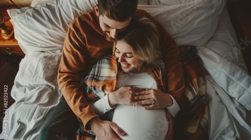 A happy young couple cuddling in bed, young woman is pregnant and her loving partner touches and caresses her belly tenderly. Top view.