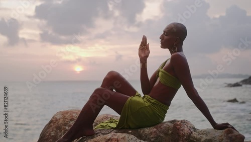 Divine non-binary black person in long open dress, brass jewelry poses on rocky hill top above dramatic ocean sunset skyline. Queer lgbtq fashion model in open outfit sits on a hill. Pantheon concept photo