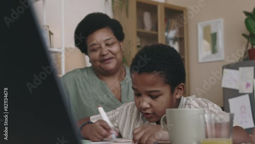 Medium of short-haired African American boy sitting at table, focused on drawing, and caring grandmother stroking him on head and encouraging, while spending time together after school