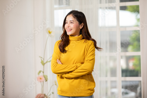 Smiling woman in yellow sweater standing with arms crossed near window in bright room with flowers, looking content and relaxed.