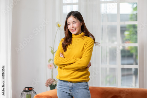 Smiling woman in yellow sweater standing with arms crossed in a cozy, bright living room with plants and an orange sofa.