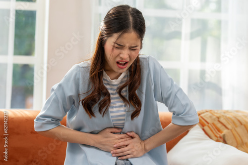 Woman experiencing stomach pain while sitting on the couch, expressing discomfort and holding her abdomen in a bright living room setting.