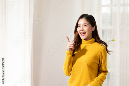 Smiling young woman in yellow sweater pointing upwards, expressing idea or inspiration, standing in brightly lit room.