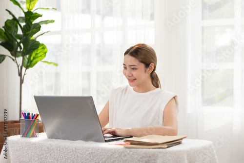 Smiling woman working on a laptop at home office, surrounded by books and plants, enjoying a bright and productive daytime environment.