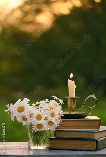chamomile flowers, candlestick with burning candle and old books on table in garden. close up. relax, reading time. romantic inspiration composition.