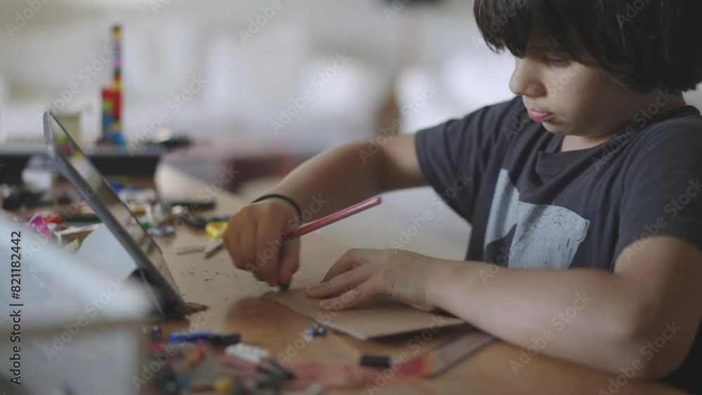 A young boy concentrates on a paper craft project while referring to instructions on a digital tablet at a table in a well-lit room. A young boy concentrates on a paper craft project while referring to instructions on a digital tablet at a table in a well-lit room.