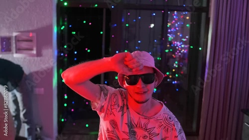 Young guy in panama hat and black sunglasses dances merrily to music, at party at night, among multi-colored flickering lights, he waves his arms and celebrates his birthday. photo