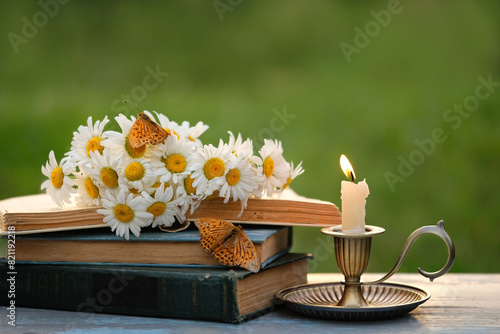 candlestick with burning candle, butterflies, old books and chamomile flowers on table in garden. close up. relax, reading time. romantic inspiration composition.