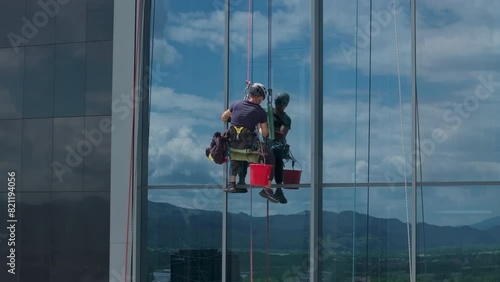 Aerial shot - High risk, dangerous job of a professional window cleaner. Window cleaner suspended on a rope cleaning window on a high rise building.