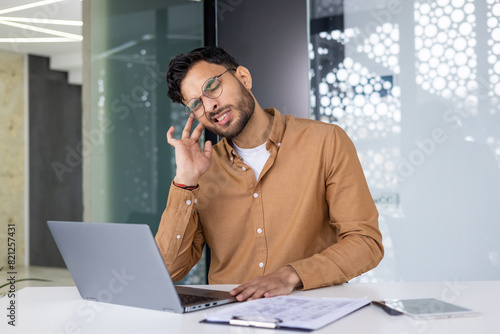 Indian young man hiding in pain, holding hand to ear, sitting at desk in modern office and suffering from inflammation