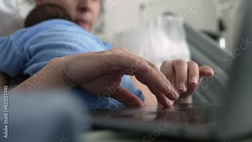Hands of new mother browsing internet on laptop while holding sleeping newborn baby, multitasking in hospital bed, combining parenting and work, postnatal care photo