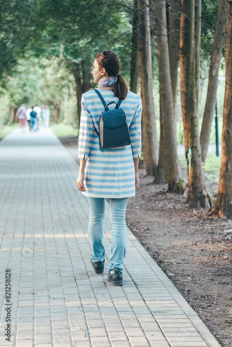 woman walks down the alley in the park, wearing jeans and a striped coat. The view from behind.