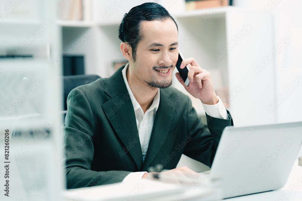 A man in a suit is talking on his cell phone while sitting at a desk with a lapt A man in a suit is talking on his cell phone while sitting at a desk with a lapt