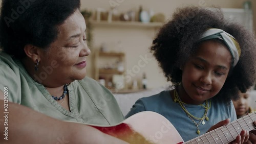 Medium of cheerful African American grandmother sharing experience, teaching curly-haired granddaughter how to play guitar, sitting on couch in homey environment