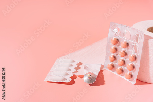 Tablets, rectal candles and ointments in packaging and toilet paper on a pink background.
