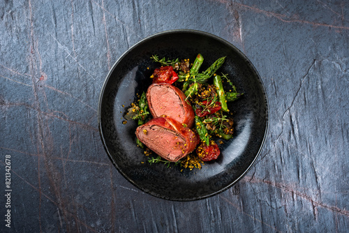 Fried mangalica pork tenderloin coated with crashed pistachio nuts, grated parmesan and chopped cranberries served with green asparagus and tomatoes as top view on a Nordic design bowl