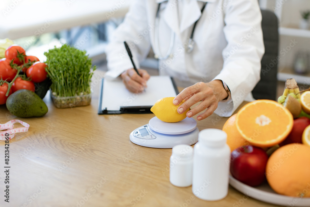Close up of hands of female nutritionist doctor sitting at table with vegetables and fruits in clinic. Positive mature Caucasian female weighing lemon while writing healthy food recipe on clipboard. Close up of hands of female nutritionist doctor sitting at table with vegetables and fruits in clinic. Positive mature Caucasian female weighing lemon while writing healthy food recipe on clipboard.