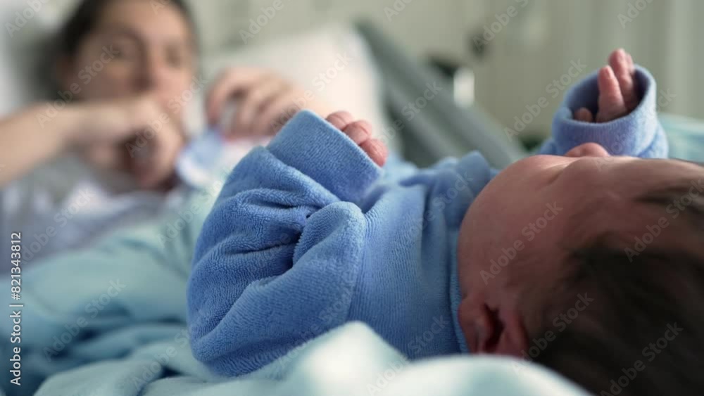 Newborn baby in a blue onesie lying on a bed in the foreground, with the mother blurred in the background in a hospital setting, highlighting the tender early moments of bonding and care Newborn baby in a blue onesie lying on a bed in the foreground, with the mother blurred in the background in a hospital setting, highlighting the tender early moments of bonding and care